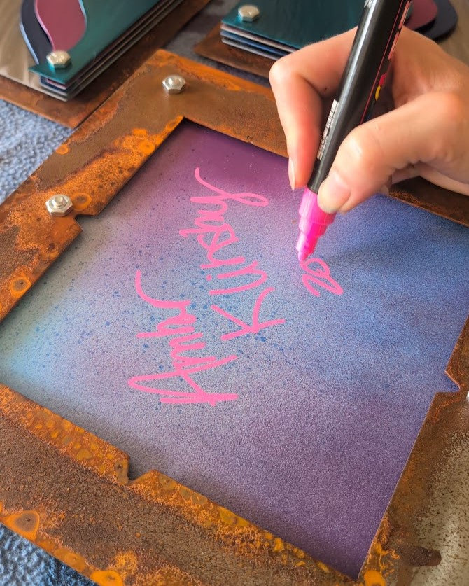 Artist Amber Klinsky signing the back of a steel panel in pink paint marker, with its rusted steel frame and other panels nearby.