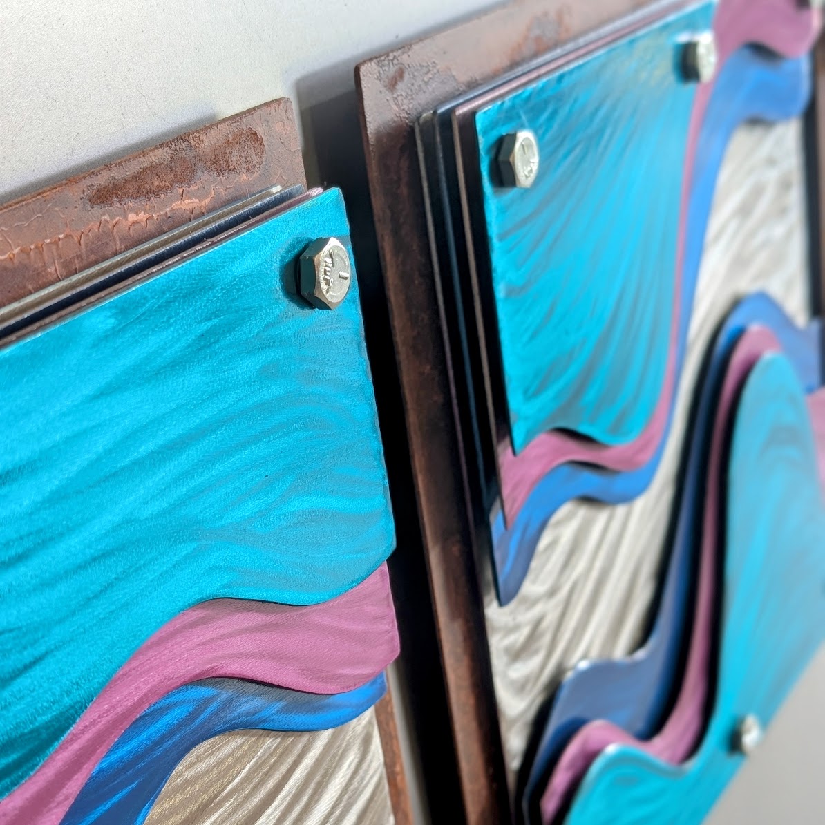 Close-up of silver hex bolts fastening teal candy-painted steel layers, with pink and blue waves and a rusted copper-toned steel frame beside them.