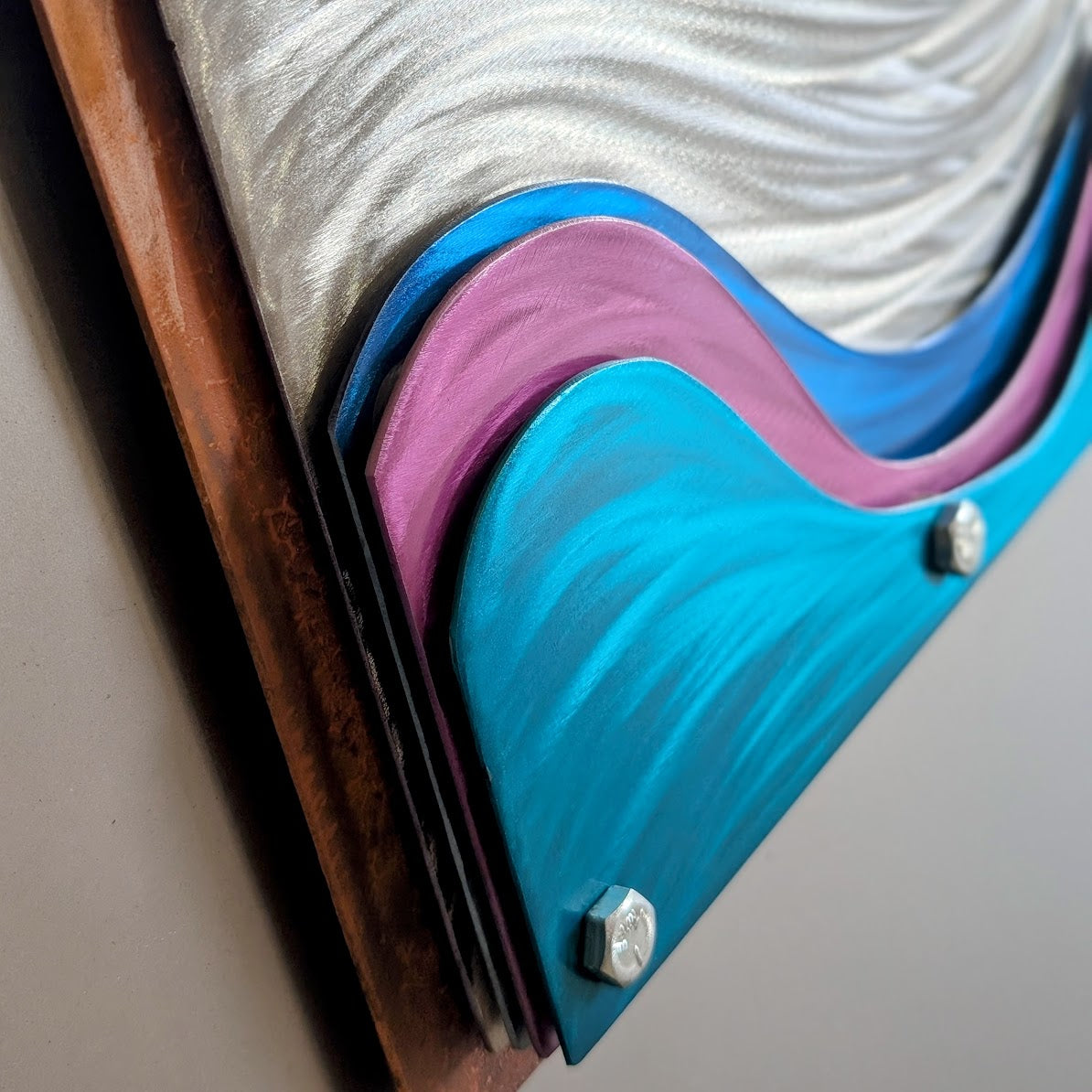 Close-up of stacked teal, pink, and blue candy-painted steel waves over swirled polished silver, showing the layered edges, a silver hex bolt, and the rusted steel frame.