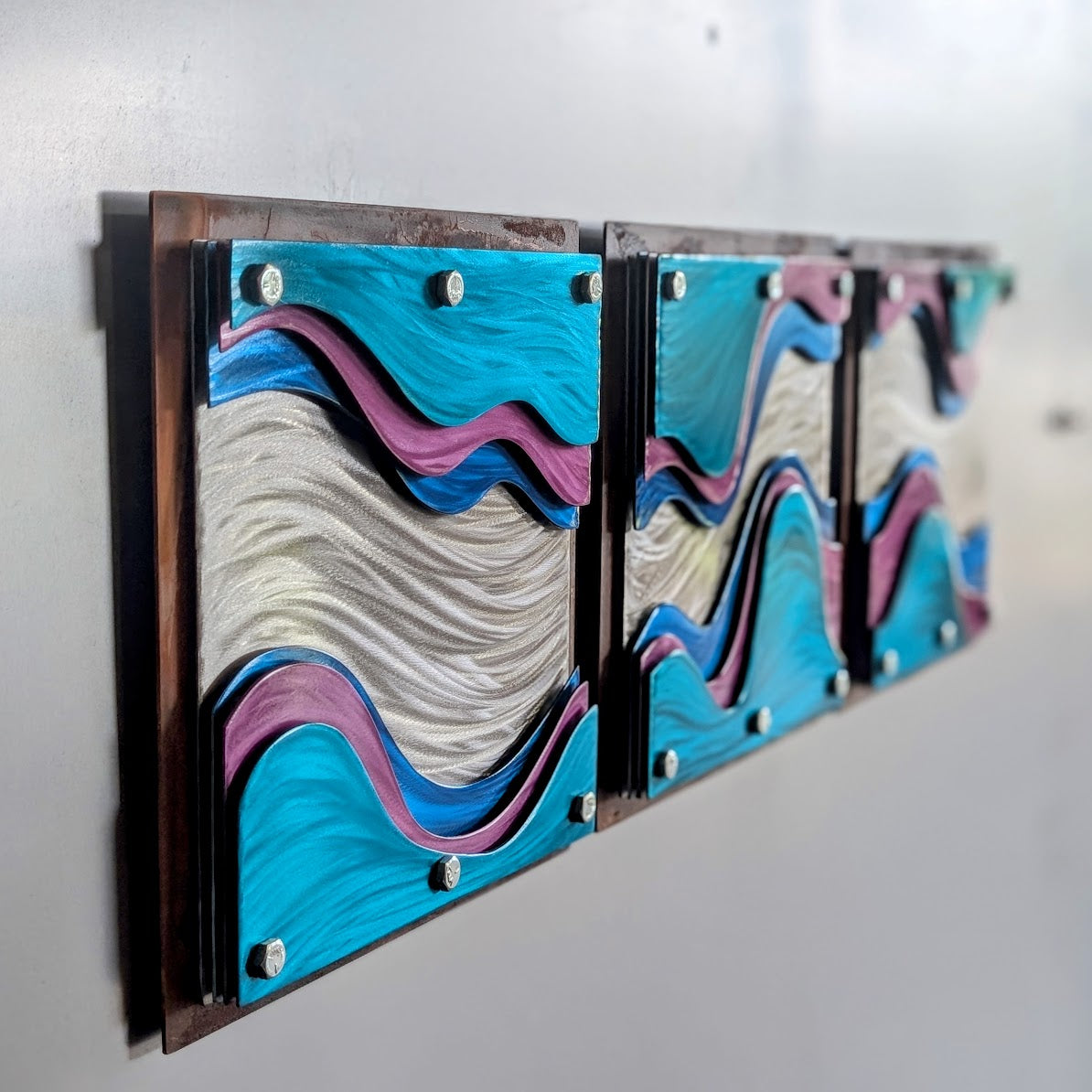 Side angle of three square layered steel panels hung in a row on a light gray wall, with candy-painted teal, blue, and pink waves around polished silver inside rusted steel frames.