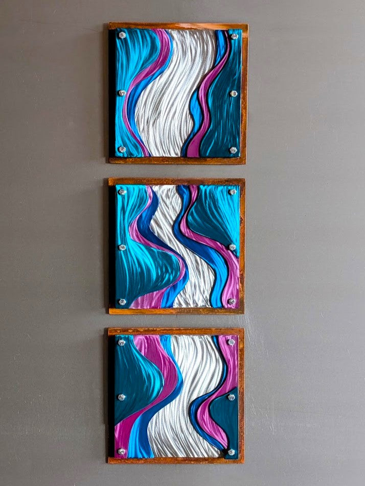 Three square steel panels stacked vertically on a gray wall, with polished silver waves winding between candy-painted teal, blue, and pink layers inside rusted steel frames.