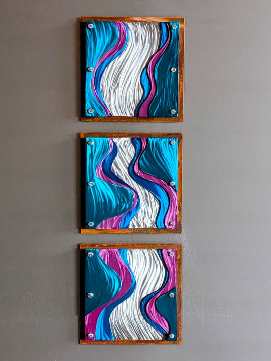 Three square steel panels stacked vertically on a gray wall, with polished silver waves winding between candy-painted teal, blue, and pink layers inside rusted steel frames.