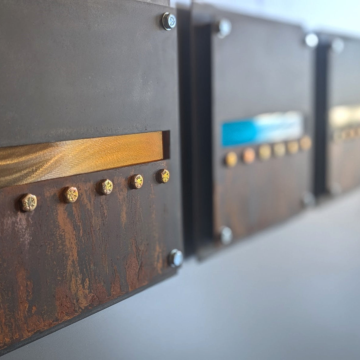 Close-up of a rusted steel panel with a polished orange candy-painted horizon band above a row of yellow zinc hex bolts, with the other two panels softly blurred behind it.
