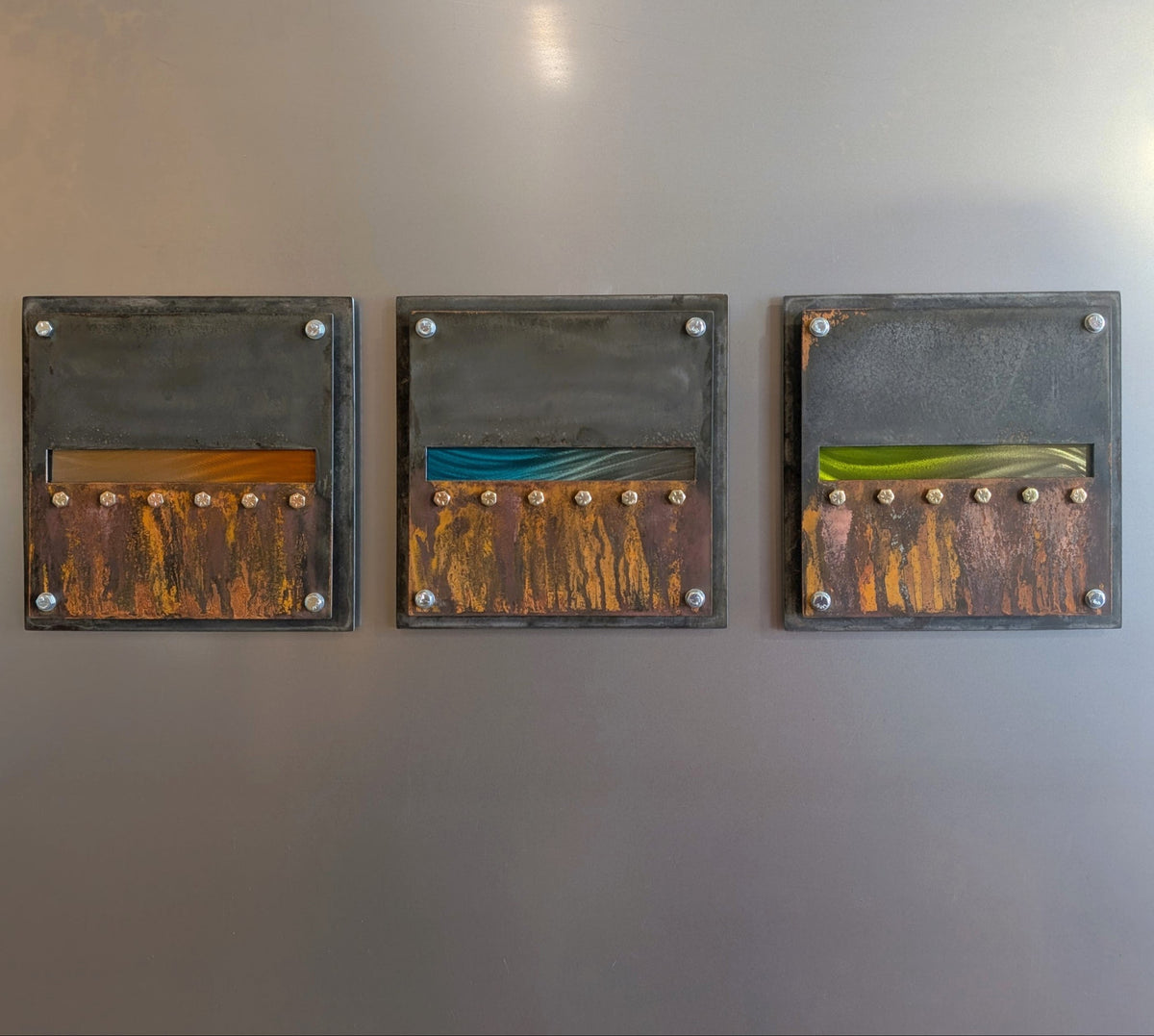 Front view of Edge of Day, three square rusted steel panels with orange, teal, and absinthe green horizon bands and exposed hex bolts.

