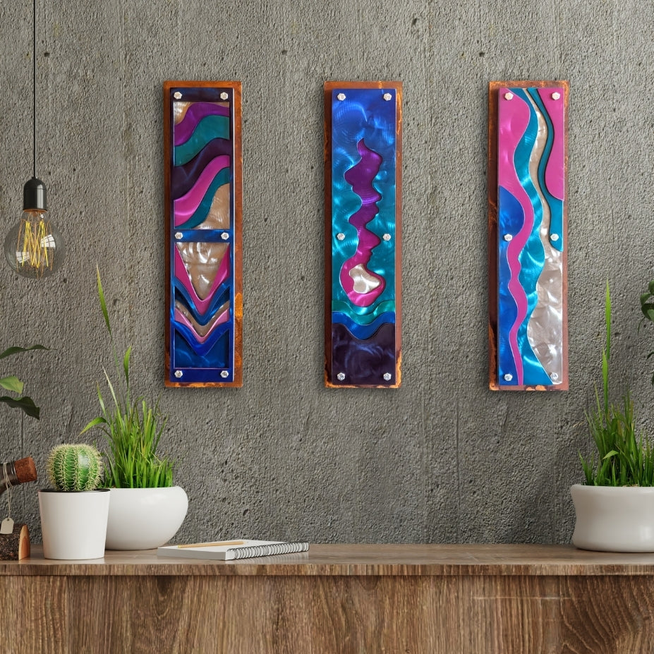 Concrete wall with plants mockup: Confluence, three layered steel wall panels in candy-painted pink, teal, purple, and blue waves, hanging side by side on a concrete wall between potted plants.