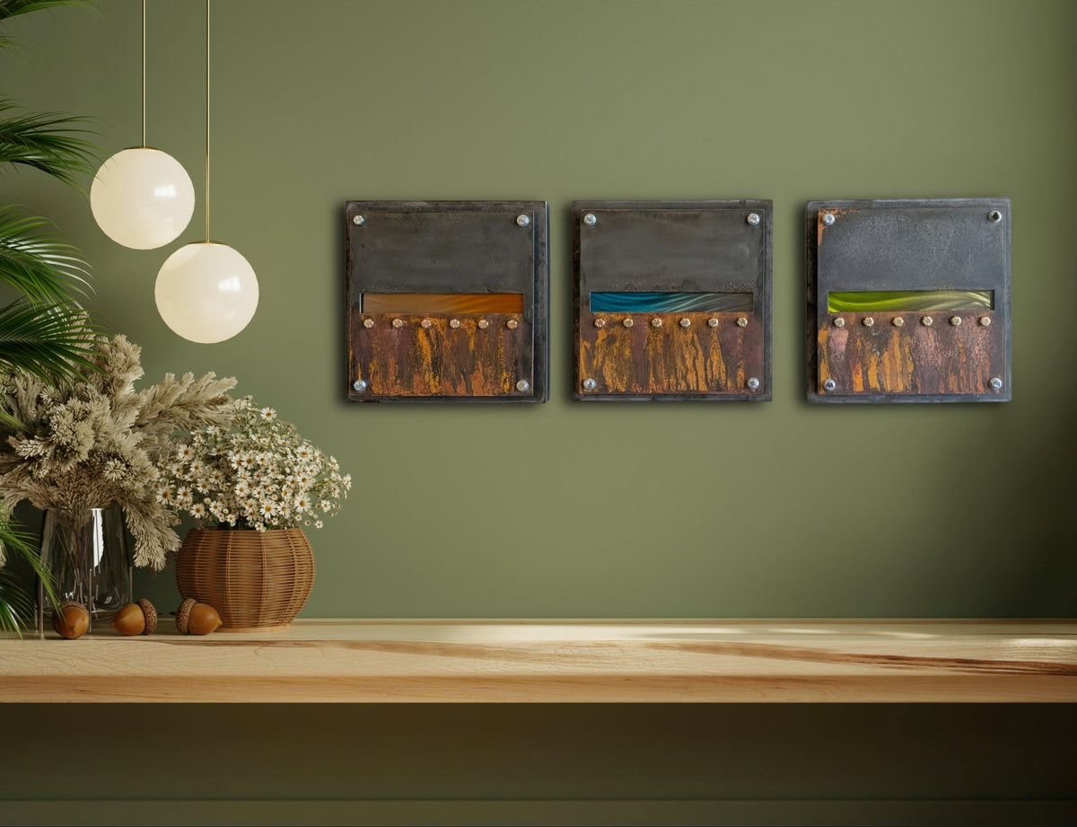 Edge of Day's three rusted steel panels displayed on a sage green wall above a wood shelf with dried flowers and pendant lights.