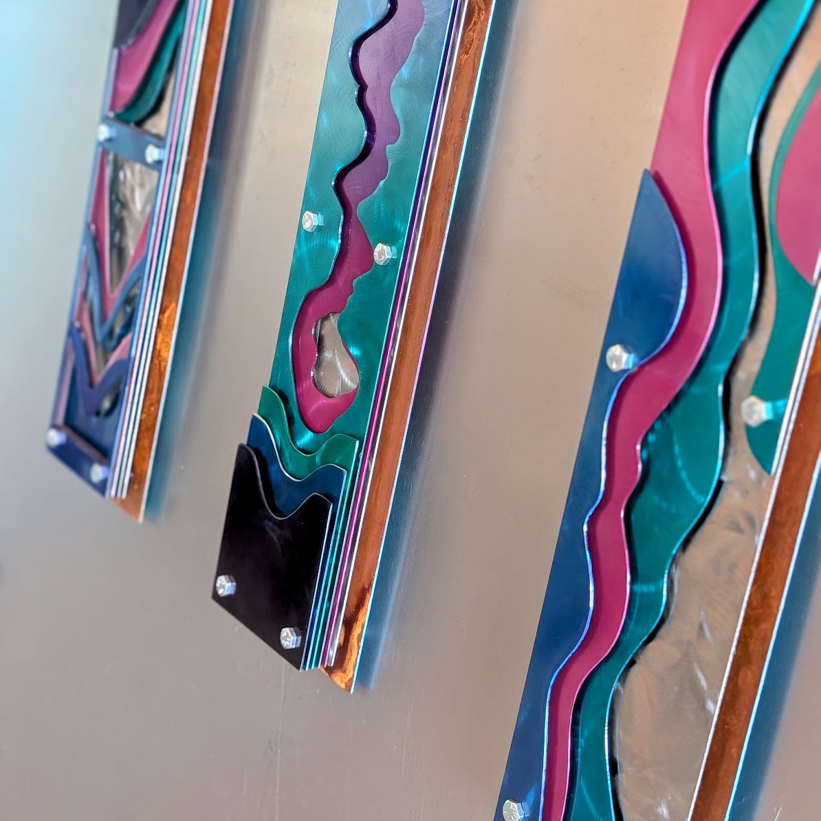 : Angled view of the three layered steel panels on a wall, showing their depth, the bolts, and the copper-toned rusted frame edges.
