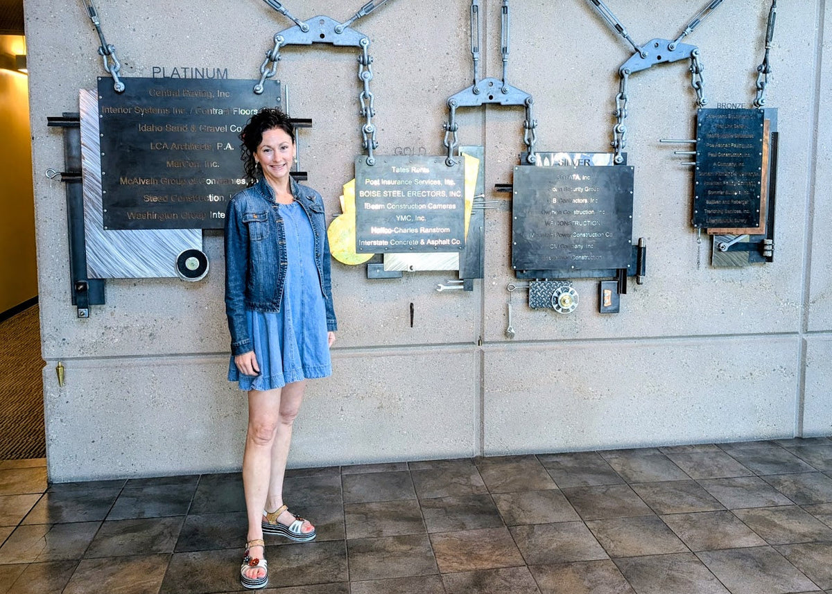 A woman wearing a blue denim jacket and a blue dress stands in front of a large industrial-style wall display with chains, plates, and plaques. The wall has 'Idaho ACC' and 'Accord Integrity Center of America' visible at the top.