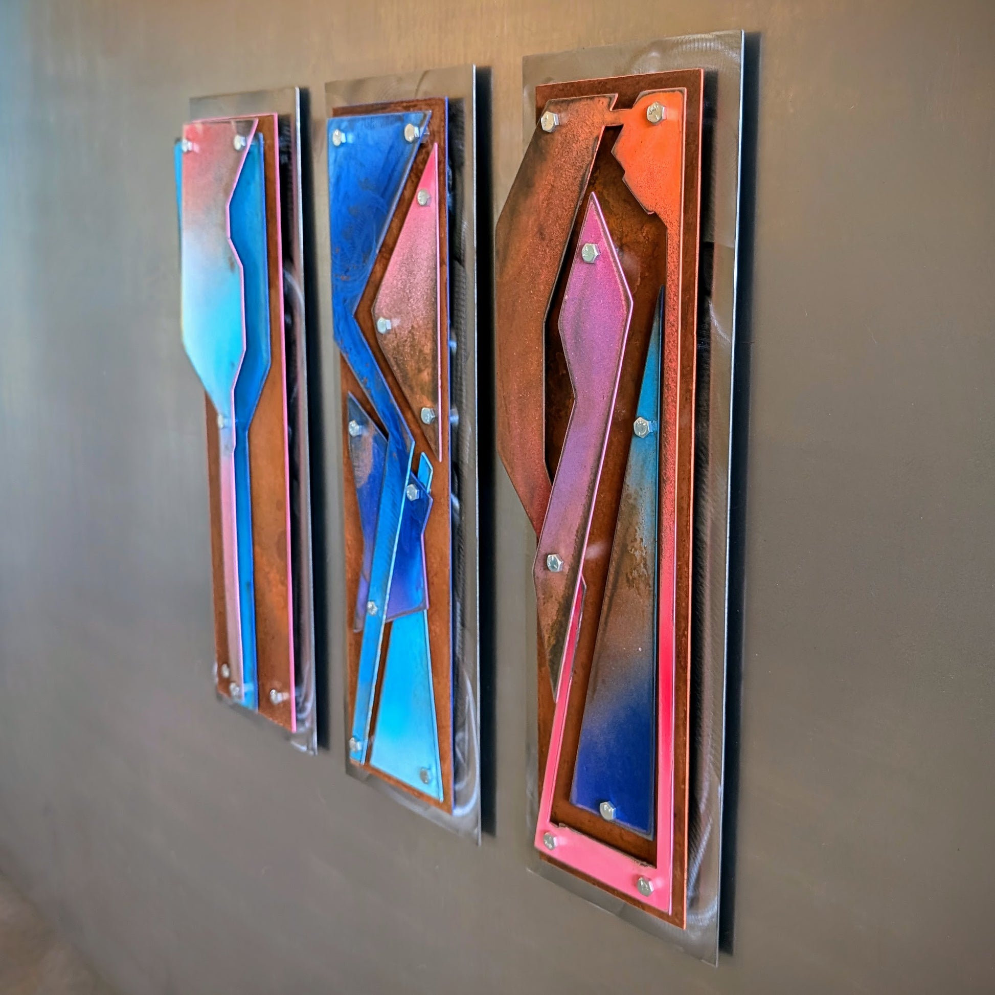 Three vertical abstract wall art panels with metallic copper, blue, pink, and orange geometric shapes, mounted on a gray wall.