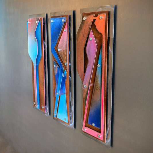 Three vertical abstract wall art panels with metallic copper, blue, pink, and orange geometric shapes, mounted on a gray wall.