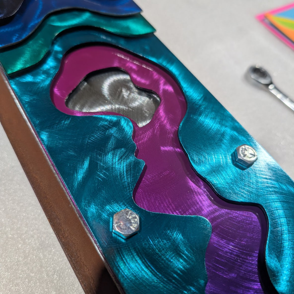 Close-up of teal, magenta, and purple candy-painted steel layers with a polished silver center and silver hex bolts.