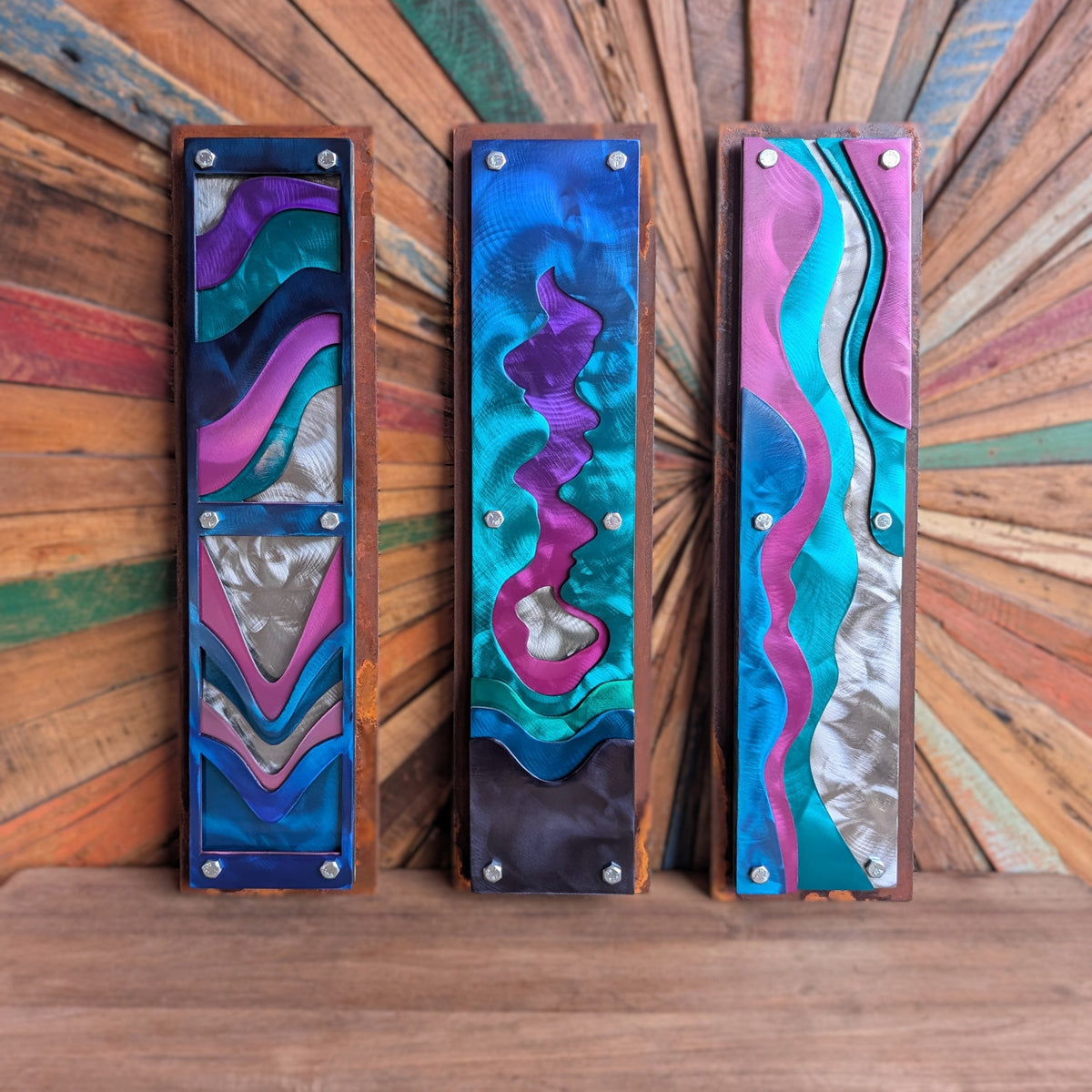 flowingwaterset: Three tall steel panels with layered wave designs in purple, teal, pink, and polished silver, bolted onto rusted steel frames, against a sunburst wood background.