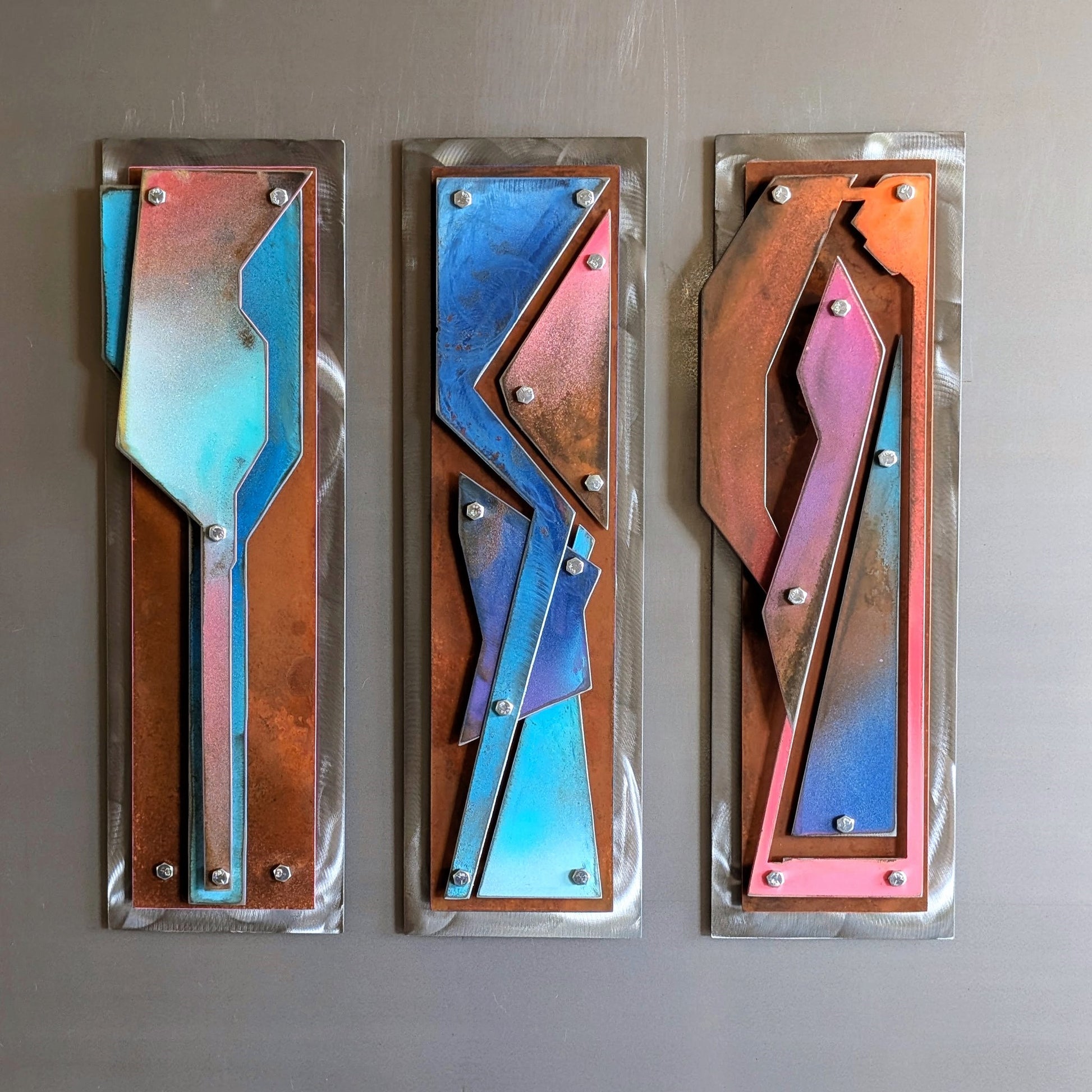 Three vertical abstract metal wall sculptures with layered geometric shapes in blue, pink, rust, and copper tones, secured by exposed hex bolts, mounted side by side on a gray wall.
