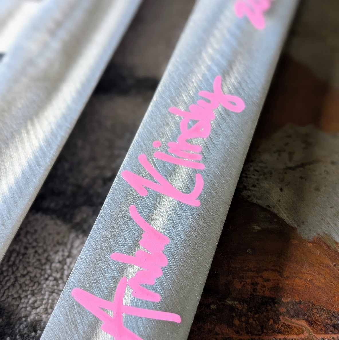 A close-up of a polished steel panel featuring the name 'Amber Klinsky' written in bright pink script, resting on a textured ruar and gray surface.